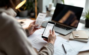 Un entrepreneur dans un petit bureau photographie un reçu avec son smartphone, avec des dossiers et un ordinateur comptable ouverts.